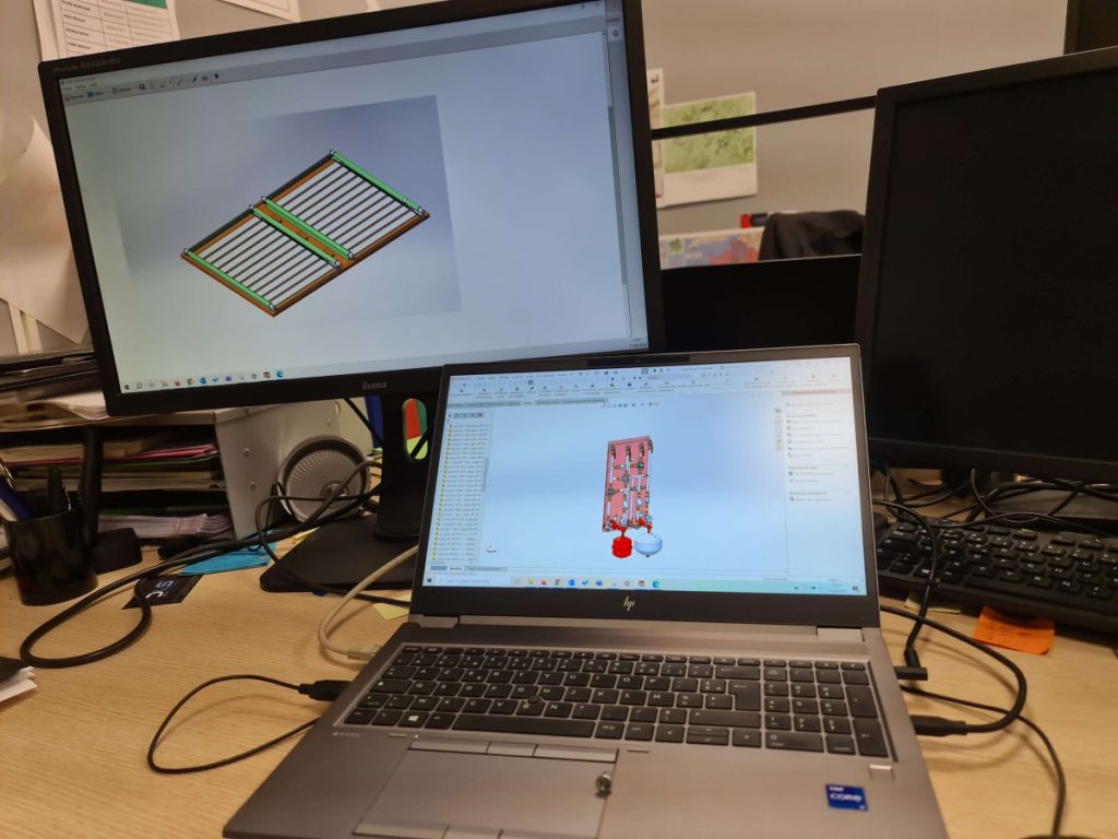 Poste de travail en bureau d’études avec ordinateur portable et écrans affichant des modèles 3D de conception mécanique sur logiciel. 