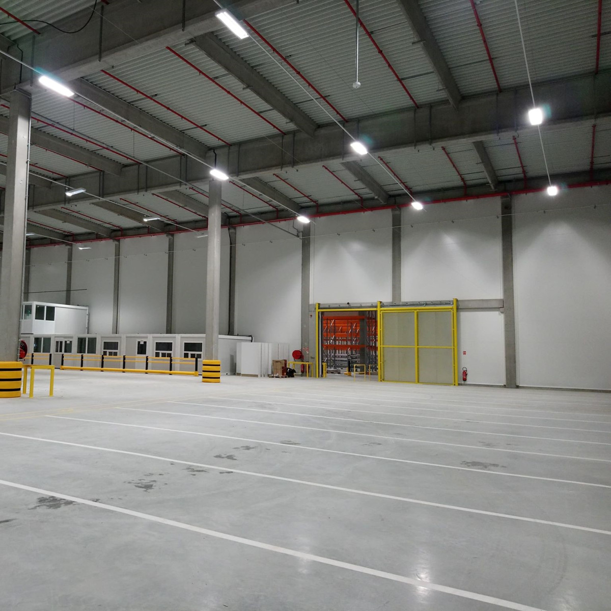 Grand entrepôt industriel vide avec sol en béton marqué de lignes blanches, hauts plafonds métalliques équipés de luminaires suspendus.