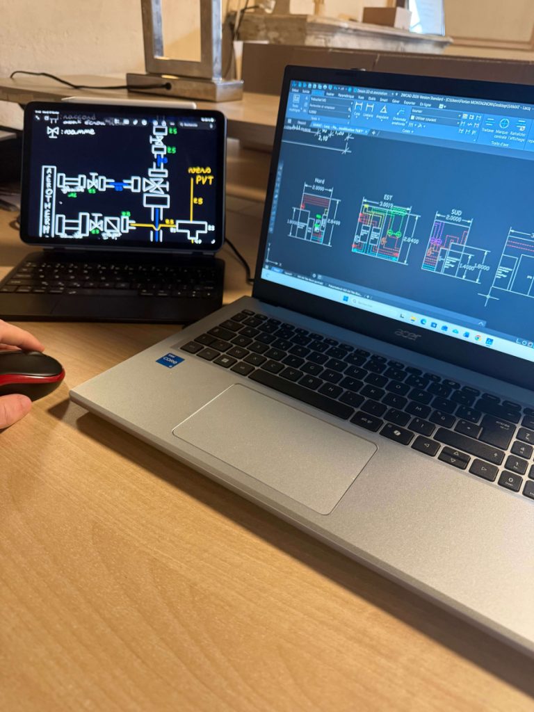 Poste de travail en bureau d’études avec ordinateur portable et écrans affichant des plans sur un logiciel. 