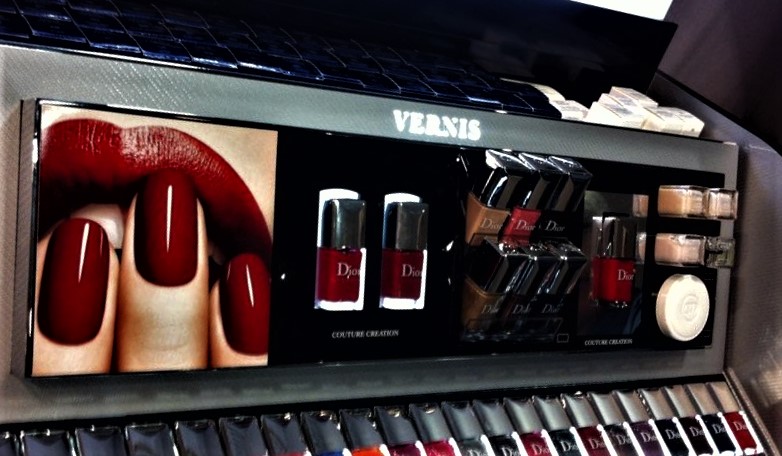 Présentation de produits cosmétiques sur un présentoir en magasin, avec mise en avant visuelle d’un produit et un visuel publicitaire en gros plan montrant des doigts aux ongles vernis rouges.