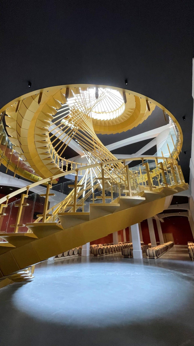 Un grand escalier en colimaçon aux caractéristiques architecturales, couleur doré avec des rampes en verre, visible à l'intérieur d'un bâtiments. 