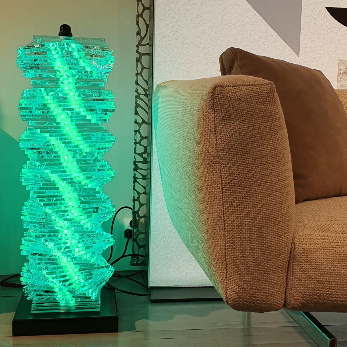 Colonne lumineuse situé sur la gauche composée de multiples couches de verre superposées, diffusant une lumière verte dans un espace lumineux à coté d'un canapé.