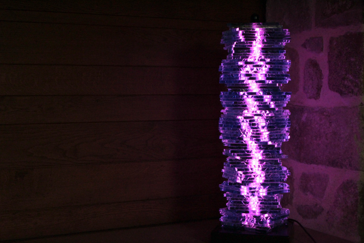 Luminaire en verre stratifié formant une colonne, éclairé par une lumière LED violette mettant en valeur la texture des plaques empilées.