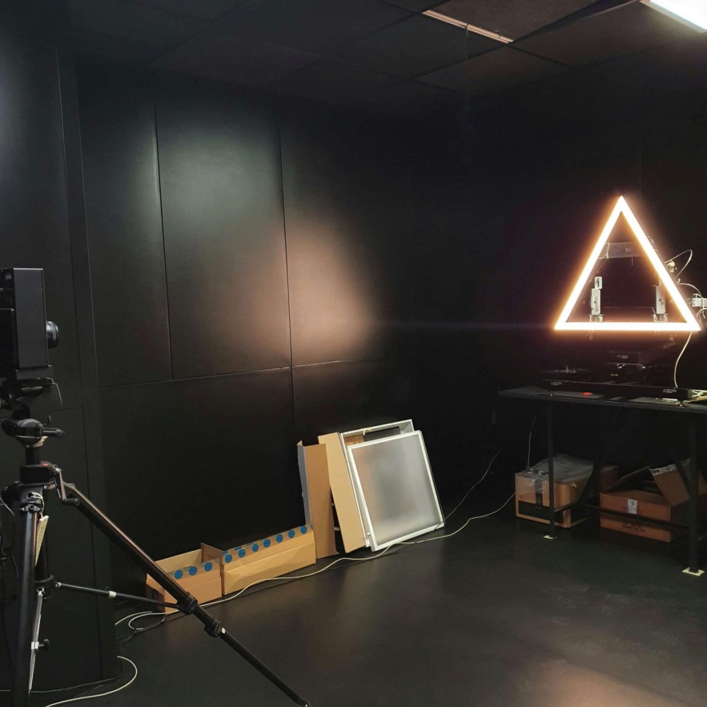 Laboratoire de test en éclairage avec murs noirs, panneau lumineux triangulaire allumé sur une table et matériel technique installé pour les essais.