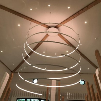 Intérieur d’une pièce avec plafond en bois apparent et poutres visibles, grand lustre circulaire moderne suspendu au centre et d’une porte vitrée donnant sur l’extérieur.