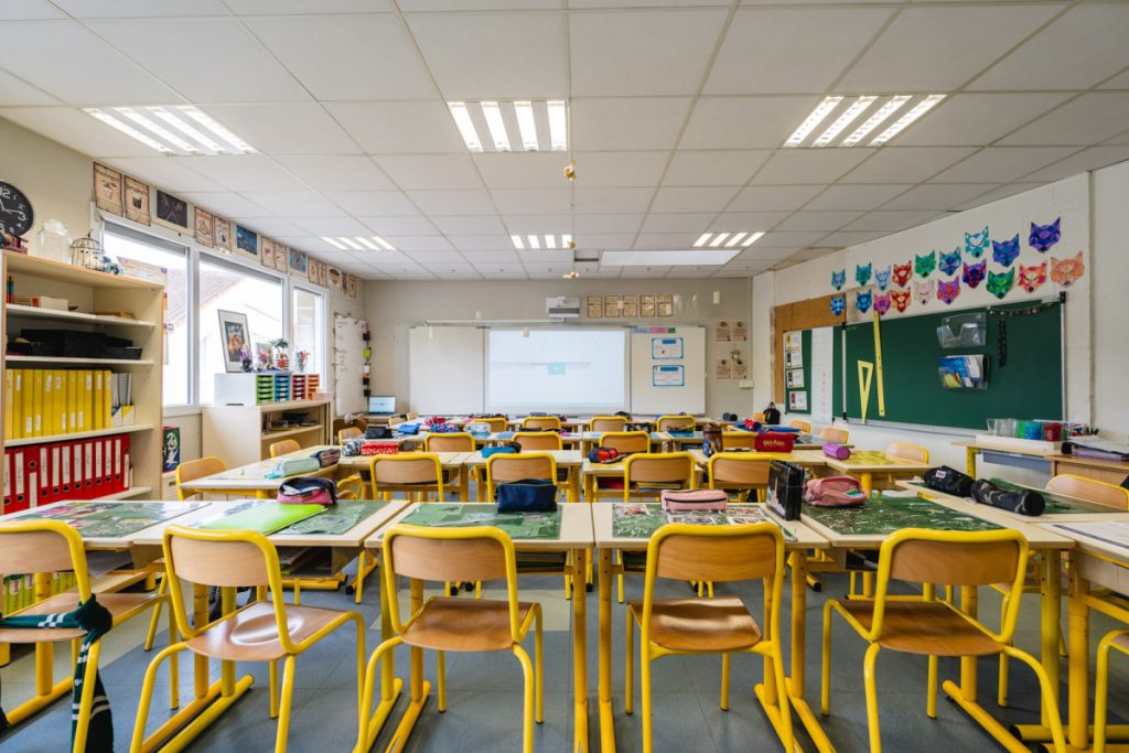 Une salle de classe vide avec plusieurs rangées de tables et de chaises jaunes, un tableau au mur et un éclairage au plafond.